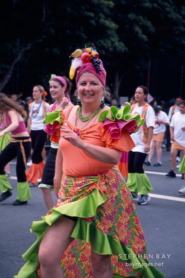 Woman dressed in orange and green wearing a hat made of fruits. Carnaval's grand parade. San Francisco.