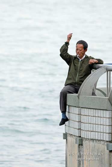 Man sitting on railing near the Hong Kong Convention Center fishing. Hong Kong, China.
